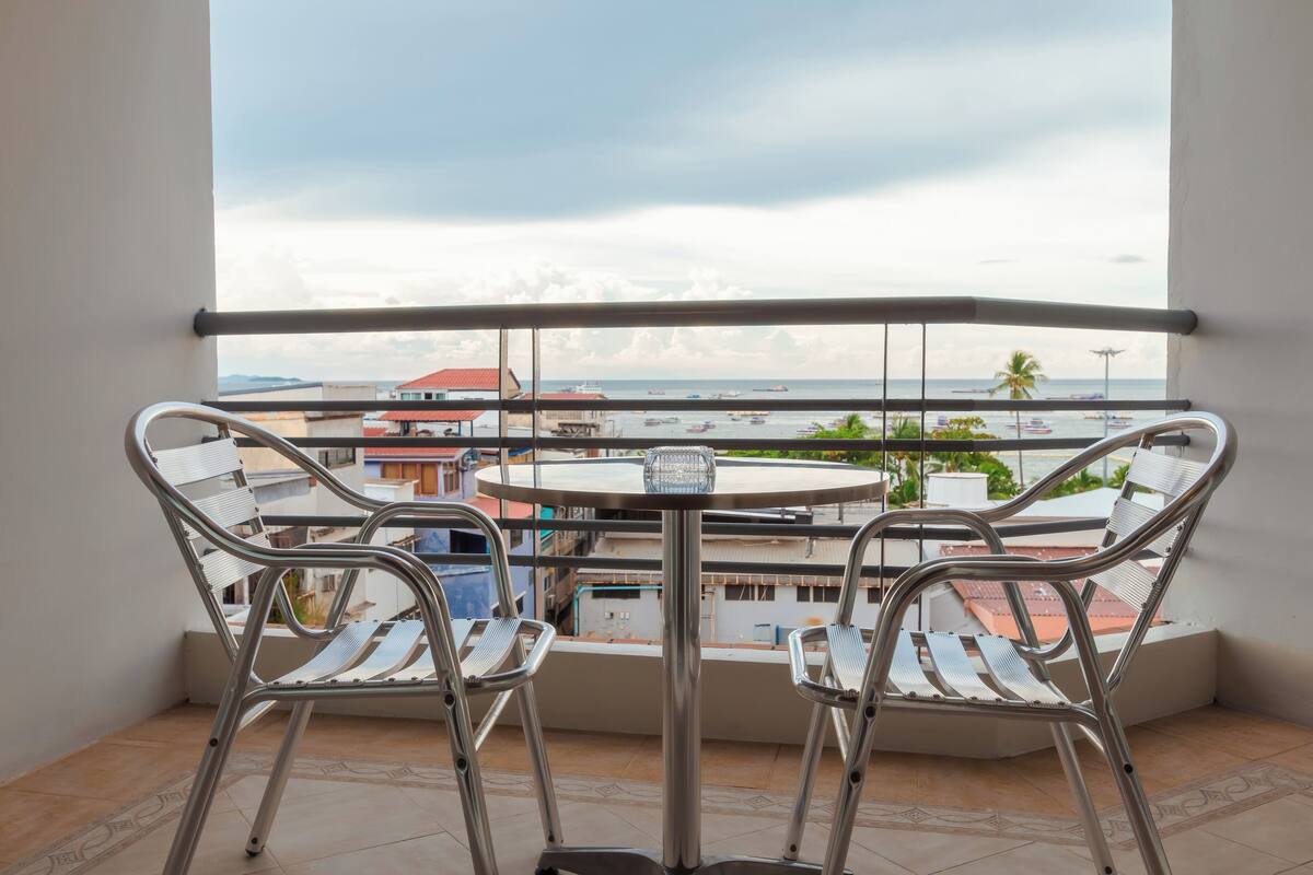 A balcony with two metal chairs and a small table, overlooking a coastal area with buildings and boats.