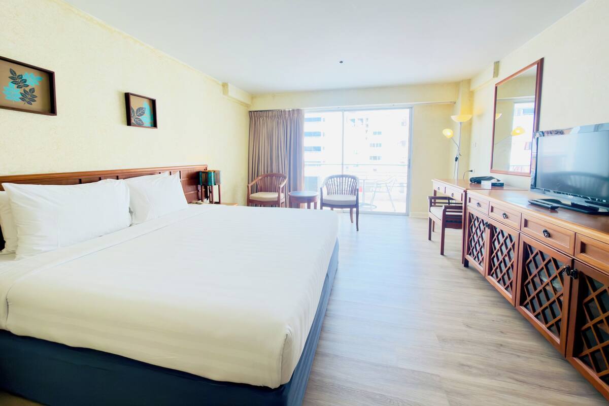 A hotel room with a large bed, a desk with a TV, a chair, and a balcony with a view of the city.