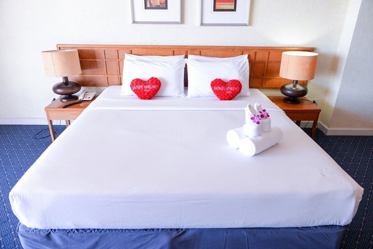 A hotel room with a large bed, two pillows with heart designs, a bedside lamp, a phone, and a towel with flowers.