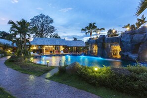 Uma área de piscina com uma cascata de pedra, espreguiçadeiras e um caminho que leva a um edifício com telhado em azulejo.