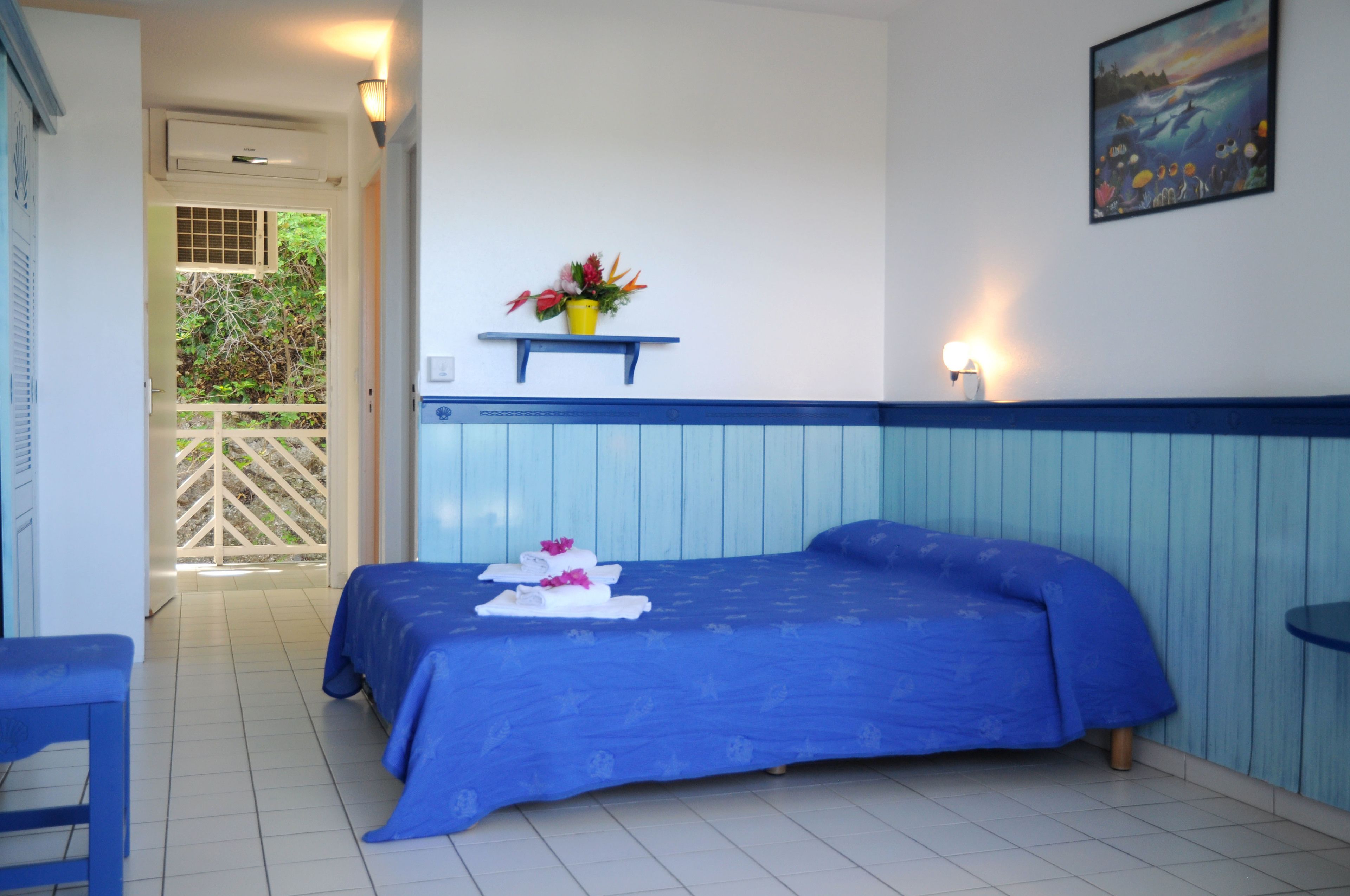 Una habitación con una cama, una mesita, un estante con un arreglo floral y un cuadro.