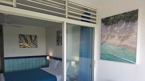 A hotel room with a bed, a table, a chair, a ceiling fan, and a painting of a beach scene. - Résidence Tropicale (Le Moule)