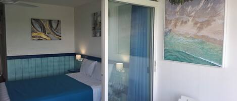 Quarto de hotel com cama, mesa, cadeira, ventilador de teto e uma pintura de uma cena de praia.