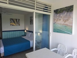 Habitación de hotel con cama, mesa, silla, ventilador de techo y una pintura de una playa.