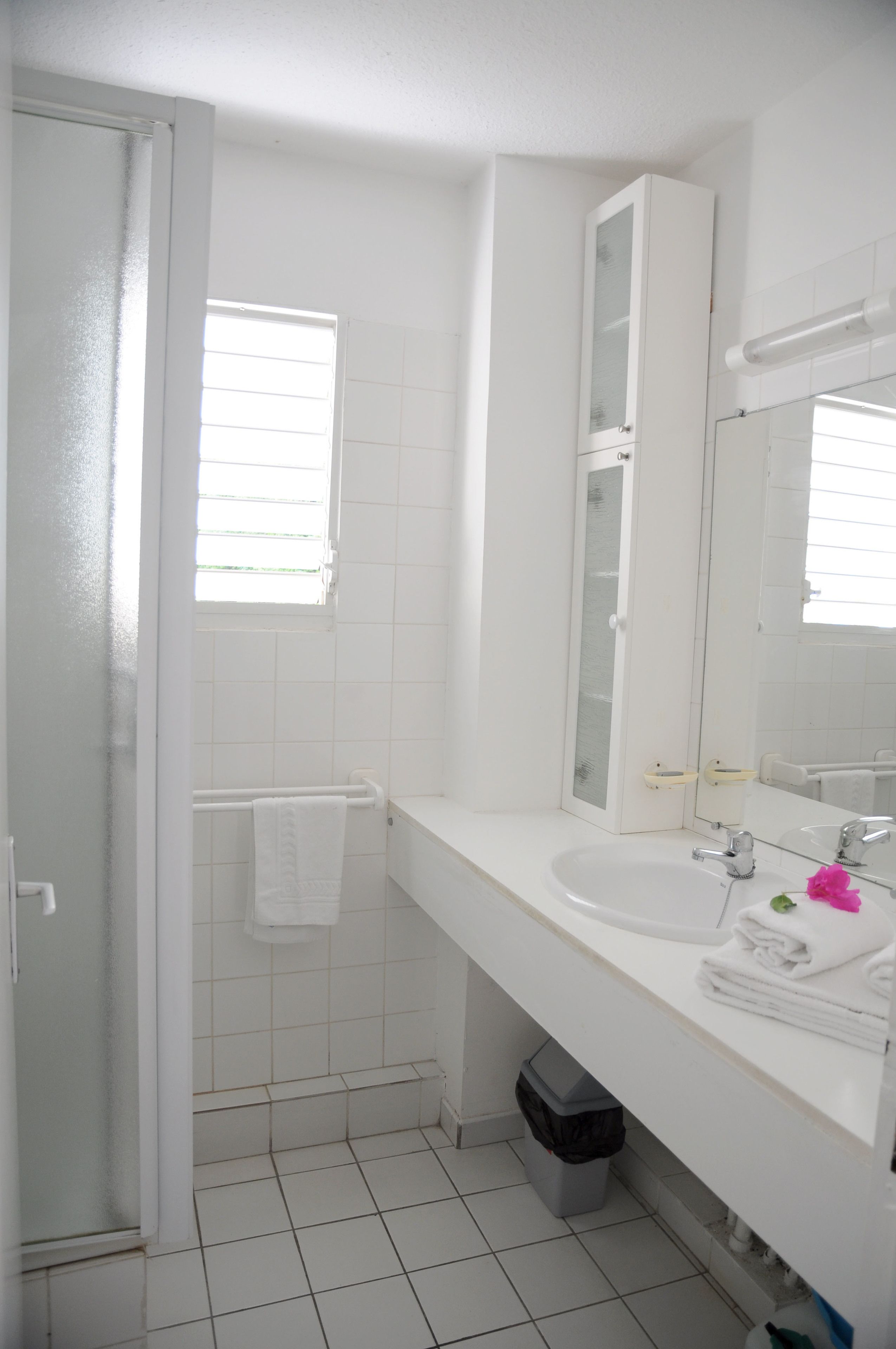 A bathroom with white tiles, a sink, a mirror, a towel rack, and a trash bin.