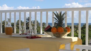 A table with a fruit bowl, a glass pitcher, and a wine glass on a balcony with a view of the ocean and palm trees. - Résidence Tropicale (Le Moule)