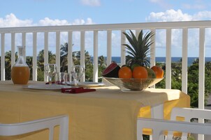 Una mesa con un frutero, una jarra de vidrio y una copa de vino en un balcón con vistas al océano y palmeras.