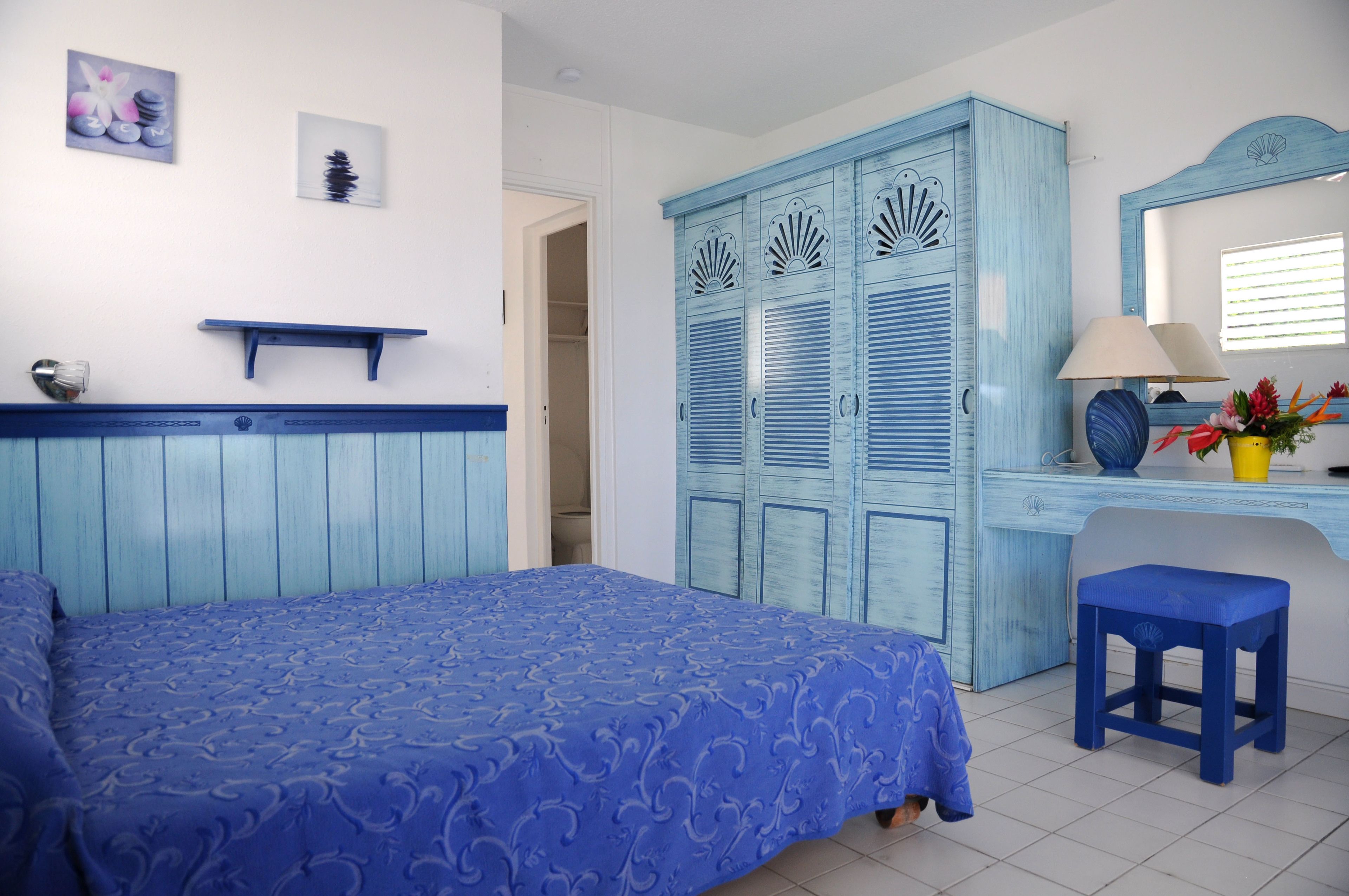 A bedroom with a bed, a blue wardrobe, a desk with a lamp, and a small stool.