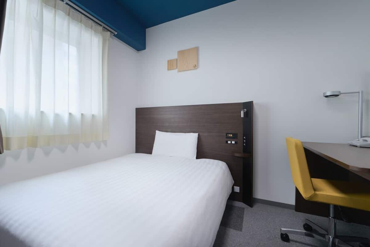 A hotel room with a bed, a desk, a chair, a lamp, and a window with curtains.