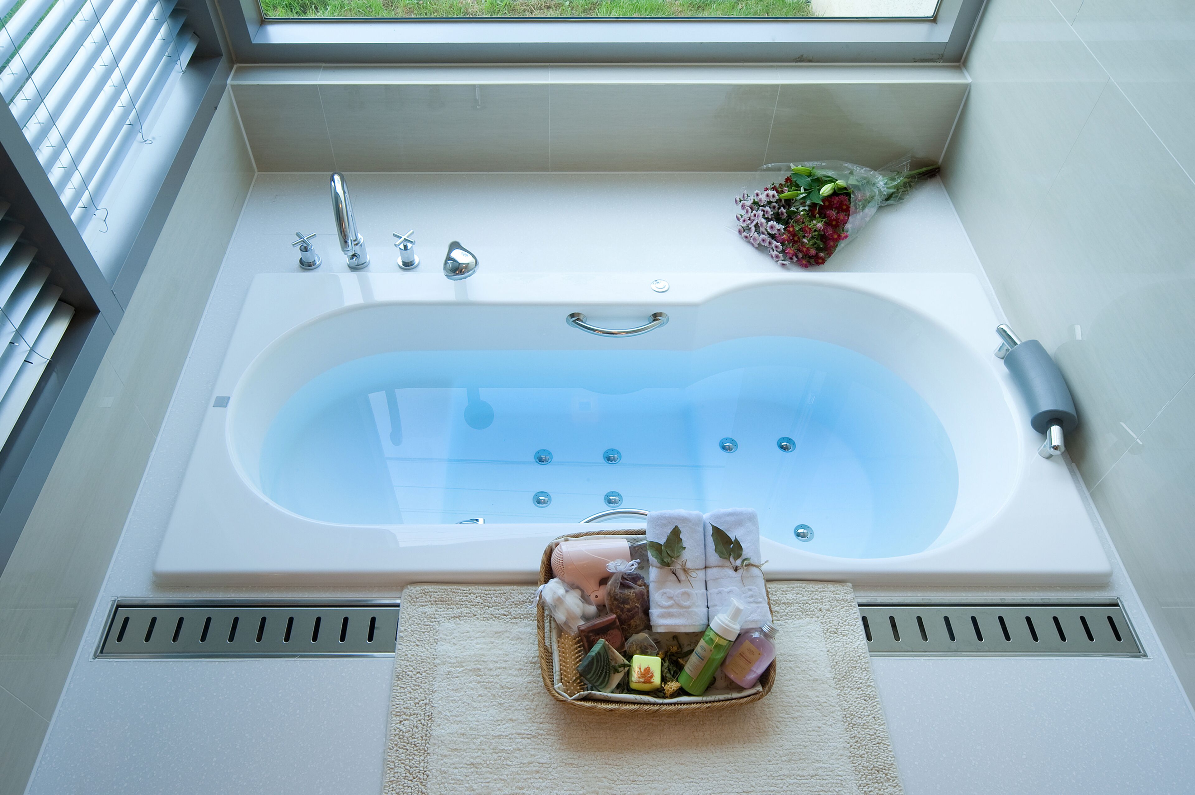 Une salle de bain moderne avec une grande baignoire, une fenêtre au-dessus et un panier contenant des articles de toilette posé sur le sol.