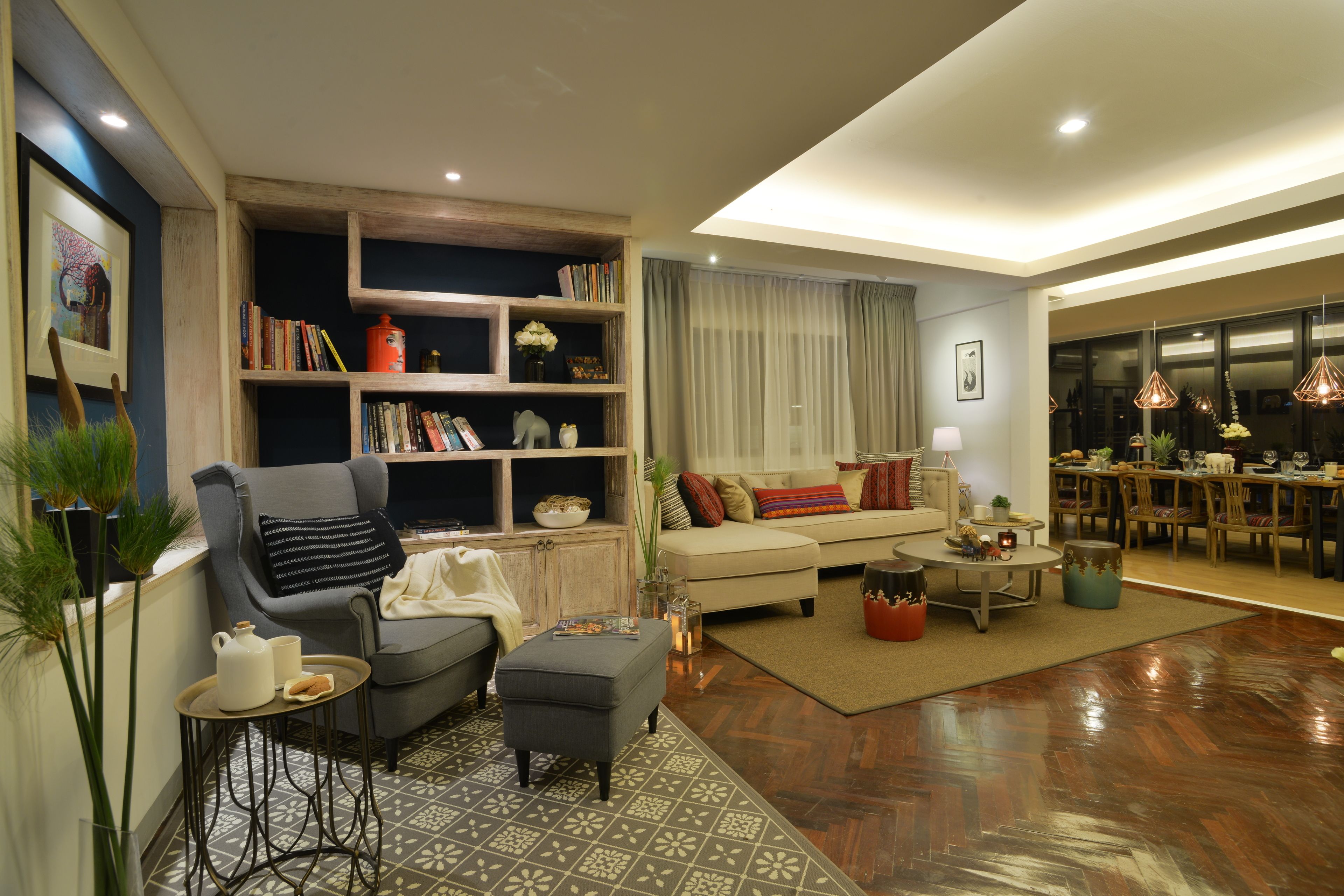 Una sala de estar moderna con sofá, sillón, reposapiés y una mesa de centro. Se observa una estantería con libros y objetos decorativos. Al fondo, un comedor con mesa y sillas.