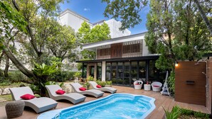 Un área de piscina moderna al aire libre con sillones de descanso y un jacuzzi, rodeada de exuberante vegetación y una casa contemporánea al fondo.