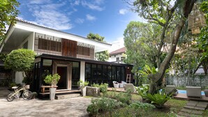 Una casa moderna con un amplio ventanal, una bicicleta y un jardín con diversas plantas y un árbol.