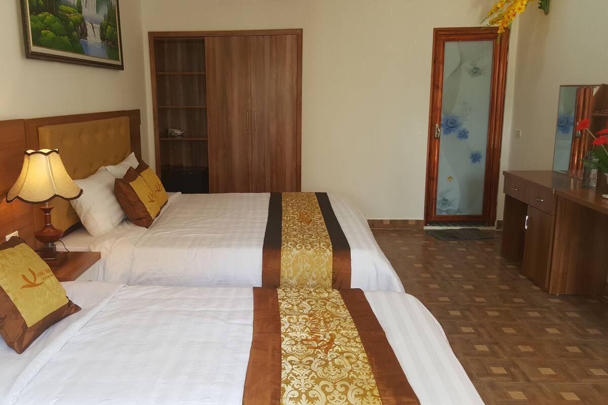 A hotel room with two beds, a wooden wardrobe, a desk with a mirror, and a painting on the wall.