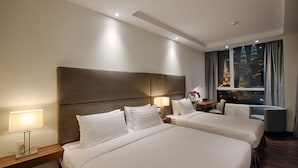 Habitación de hotel con dos camas, una mesita de noche, un escritorio, una silla y una ventana con vista a la ciudad.