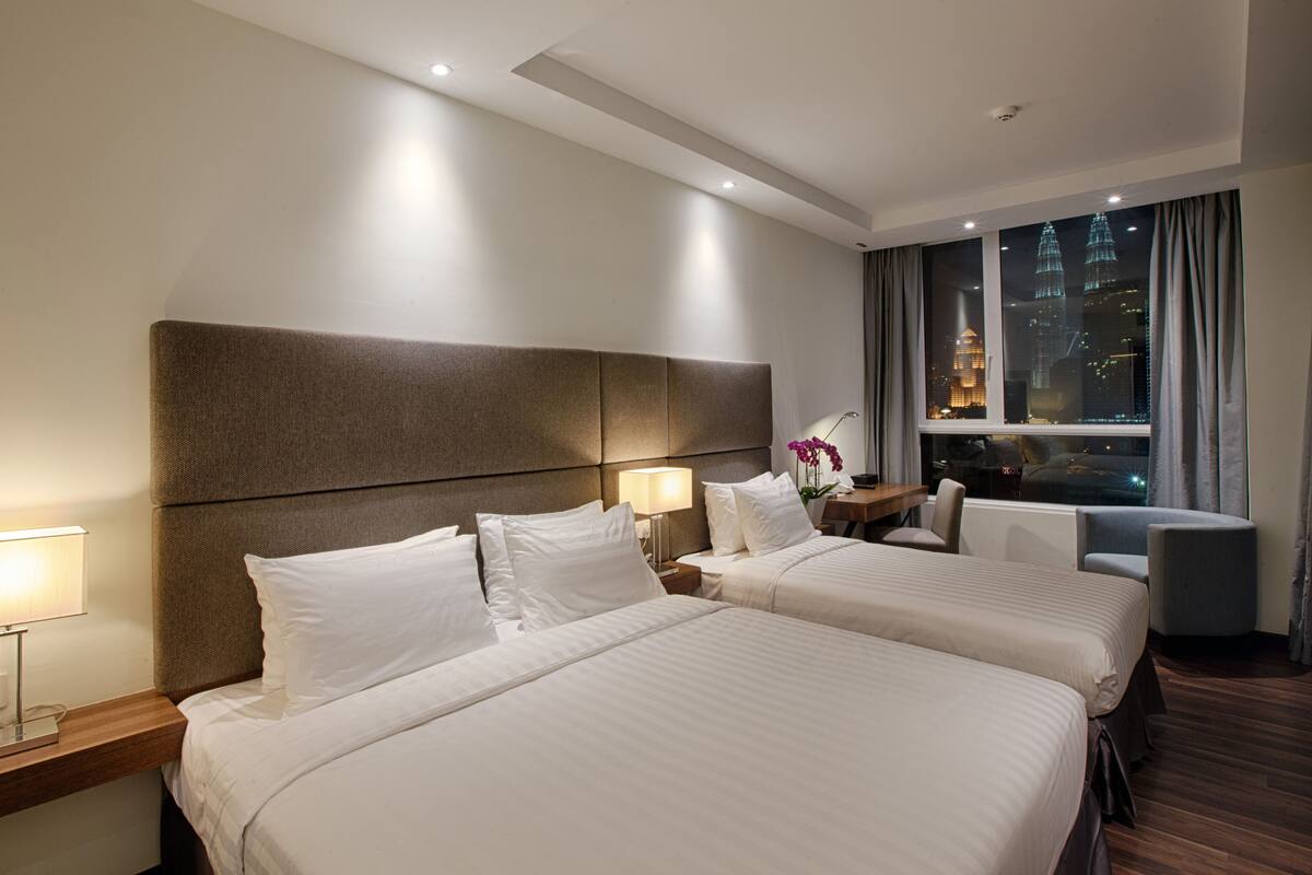 A hotel room with two beds, a nightstand, a desk, a chair, and a window with a city view.