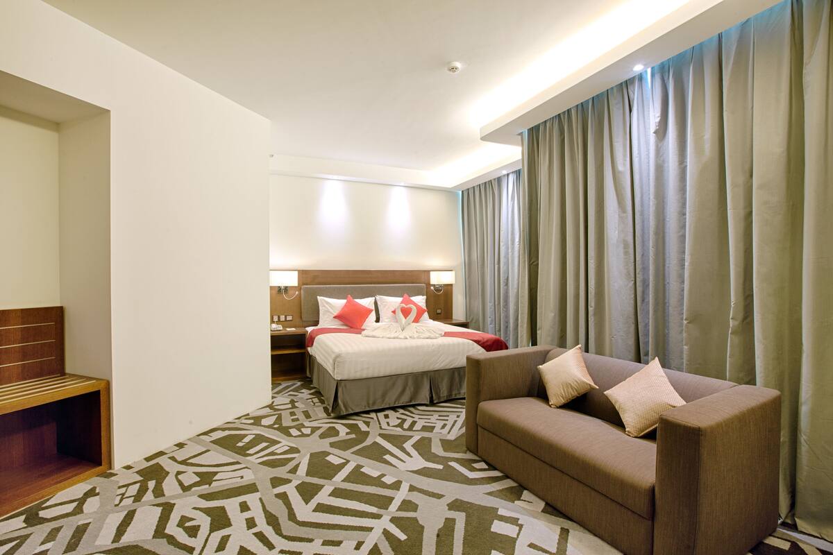 A hotel room with a bed, a sofa, a patterned rug, and a large window with curtains.