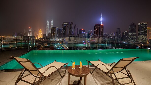 Una piscina en la azotea con sillones de descanso y una mesa, con vistas a un paisaje urbano con rascacielos iluminados.