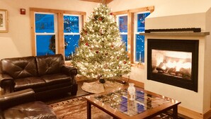 Una sala de estar acogedora con un árbol de Navidad, un sofá de cuero, una mesa de centro de vidrio, chimenea y ventanas amplias.