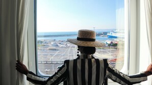 Pessoa vestindo camisa listrada e chapéu de palha, parada perto de uma janela com vista para um aeroporto.