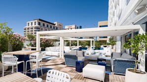 Uma área de estar ao ar livre com piso de madeira, mobiliário azul e branco e uma pérgola branca.