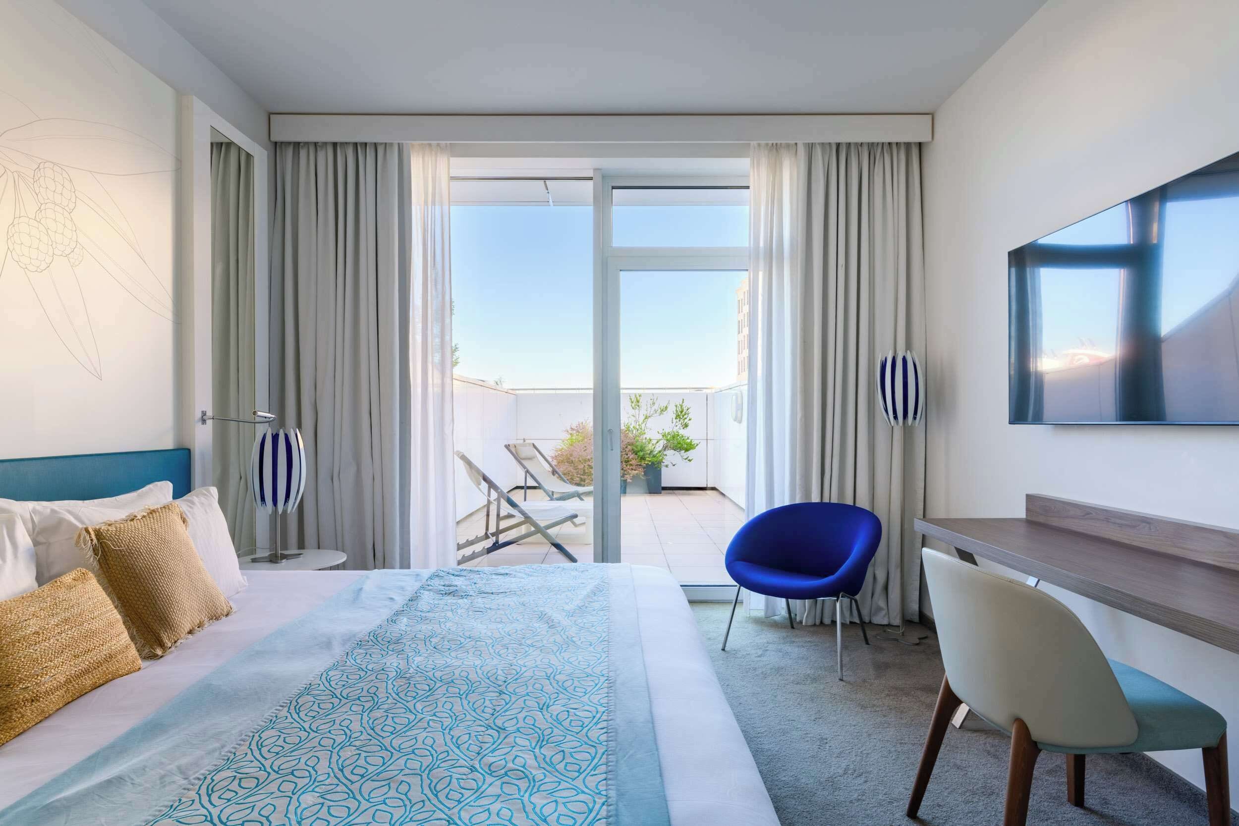 Habitación de hotel con cama, escritorio, silla, televisión y balcón con vistas.