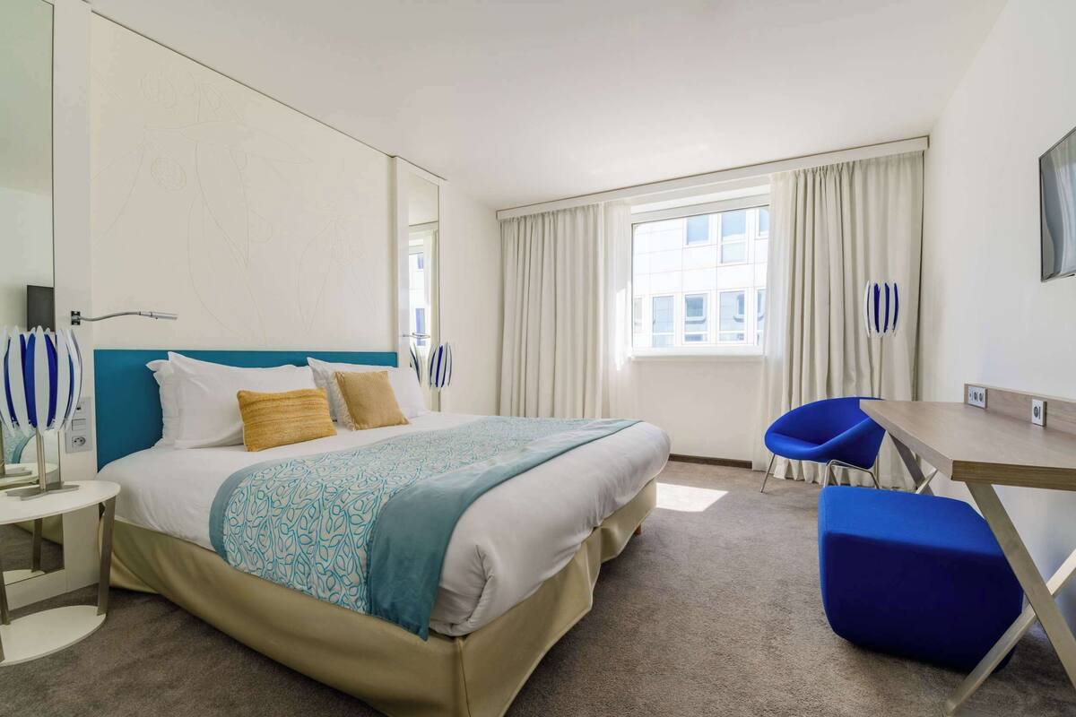 A hotel room with a bed, a desk, a chair, and a large window with curtains.