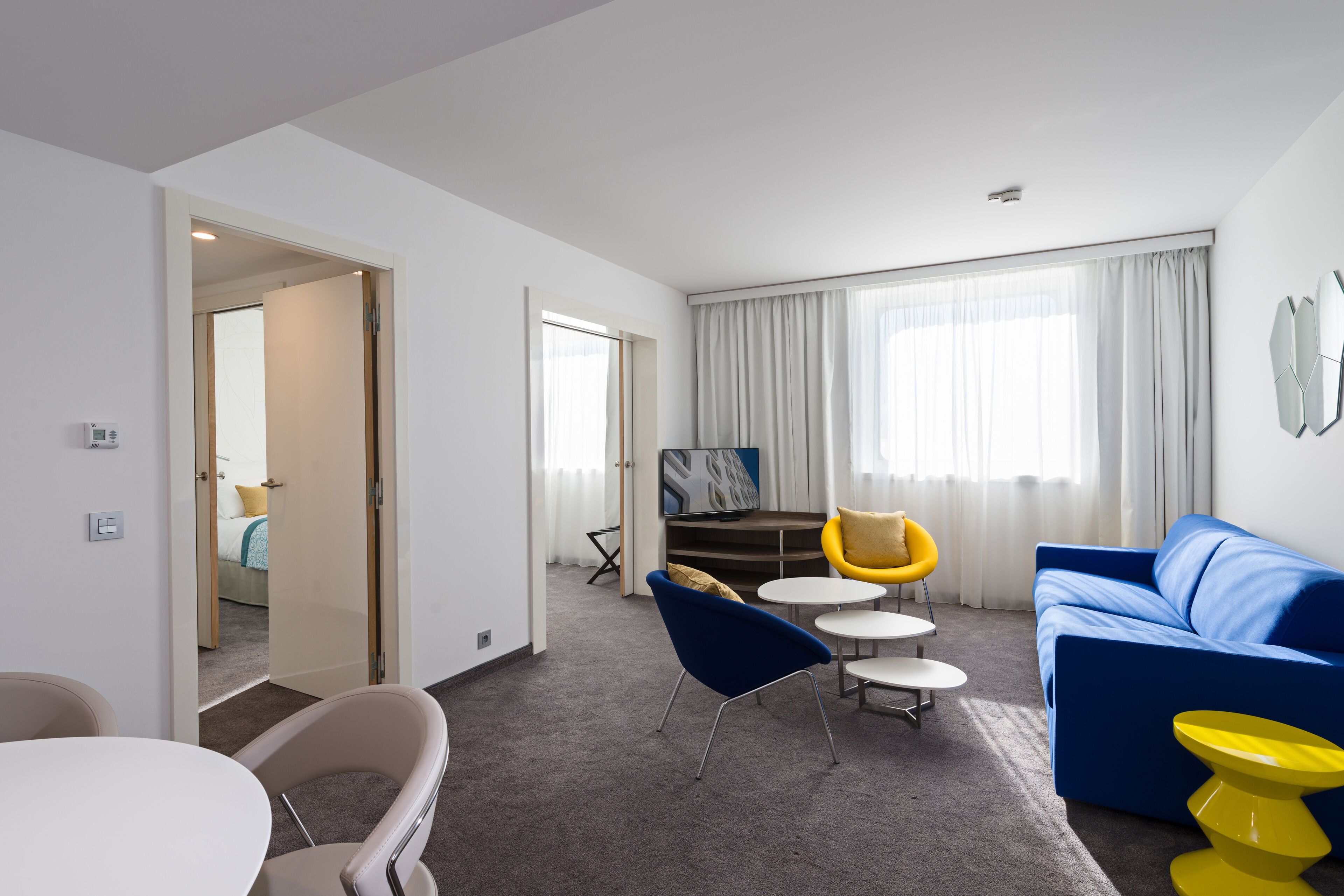 Une chambre d’hôtel moderne avec un canapé bleu, des chaises jaunes, une table blanche et une grande fenêtre avec des rideaux.