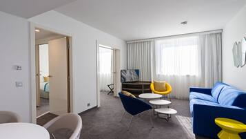 Une chambre d’hôtel moderne avec un canapé bleu, des chaises jaunes, une table blanche et une grande fenêtre avec des rideaux.