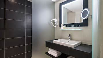 A modern bathroom with a dark tiled wall, a white sink with a mirror above it, and a shelf with towels.