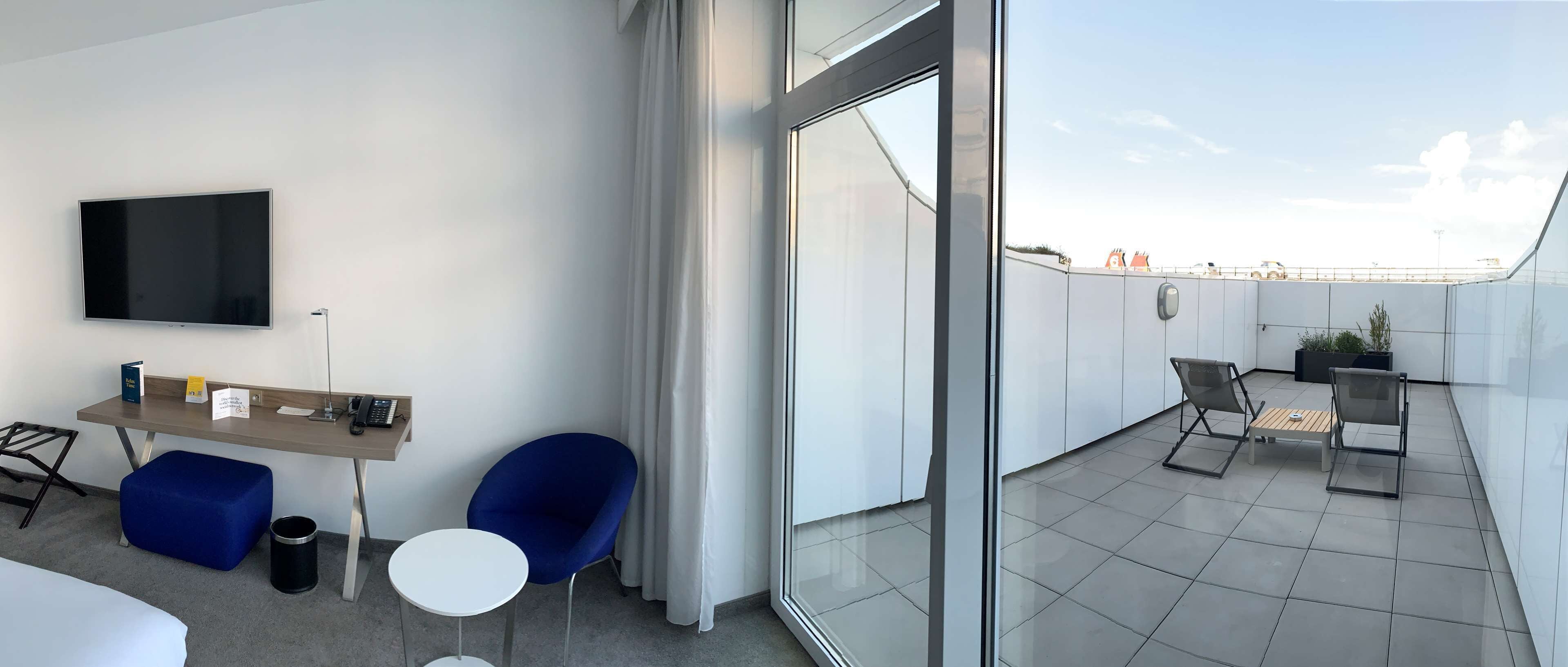 A modern hotel room with a flat-screen TV, a desk with a telephone, a blue chair, a small round table, and a large window offering a view of the cityscape.