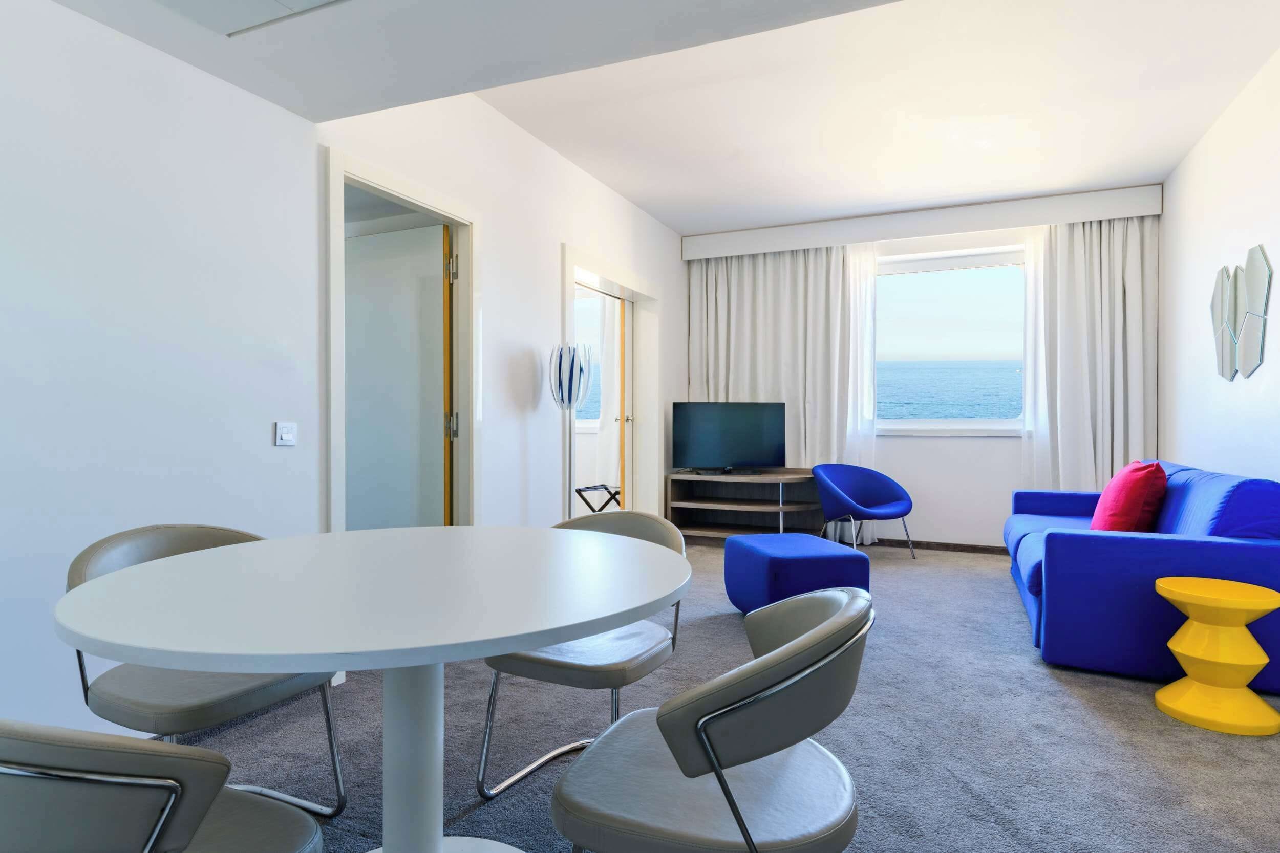 A hotel room with a round dining table, two chairs, a blue sofa, a television, and a view of the sea through large windows.