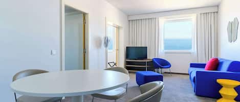 Habitación de hotel con una mesa de comedor redonda, dos sillas, un sofá azul, un televisor y vista al mar a través de amplias ventanas.
