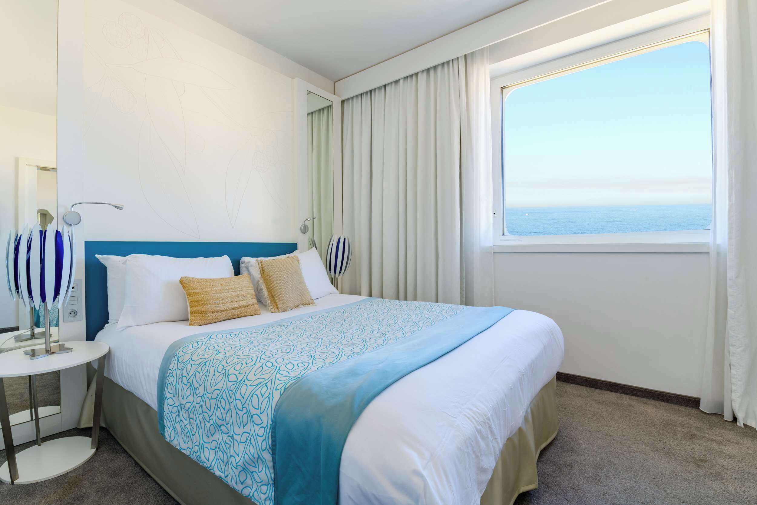 Een hotelkamer met een groot bed, een nachtkastje, een lamp en een raam met uitzicht op zee.