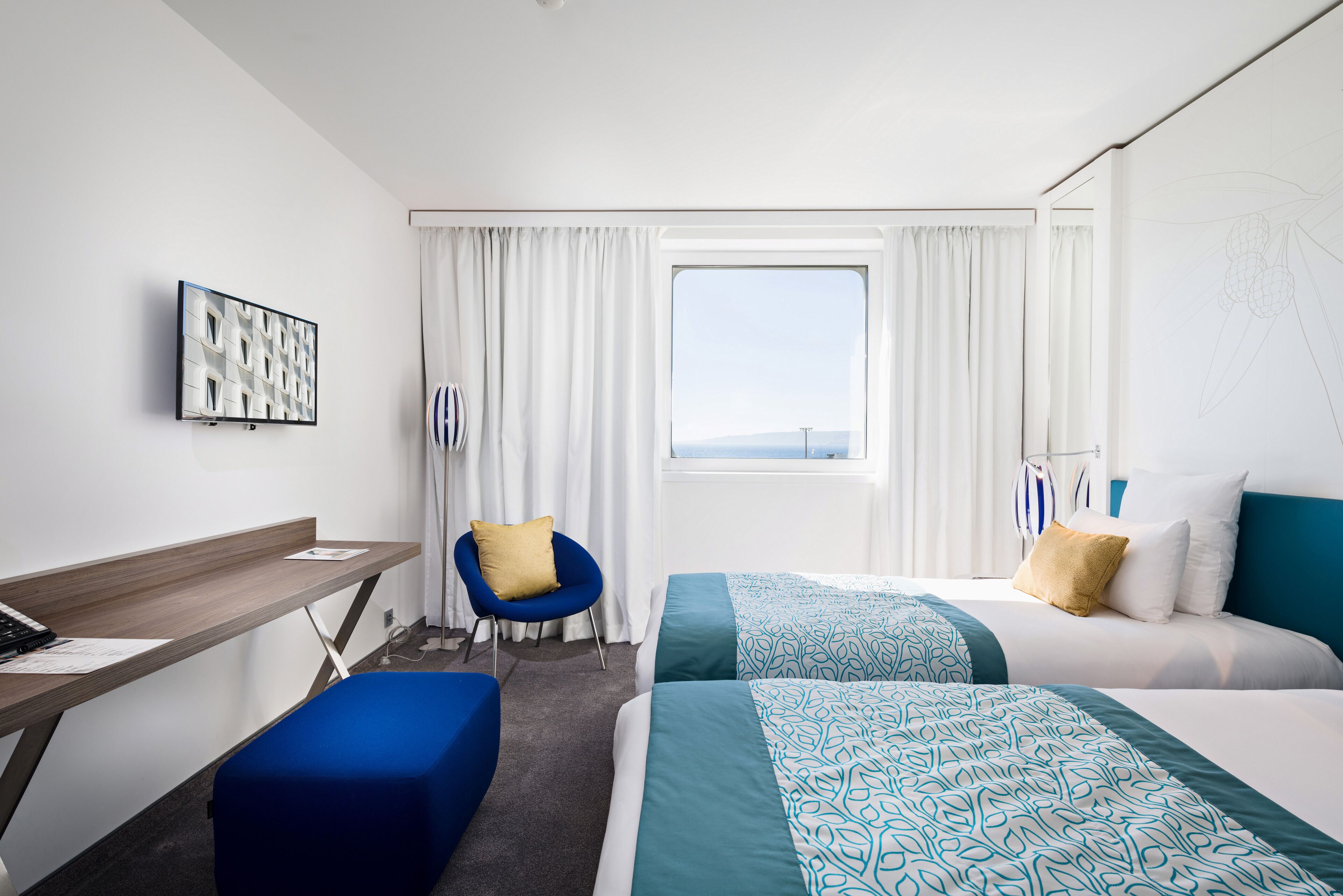 A hotel room with a desk, two beds, a chair, and a window with curtains.