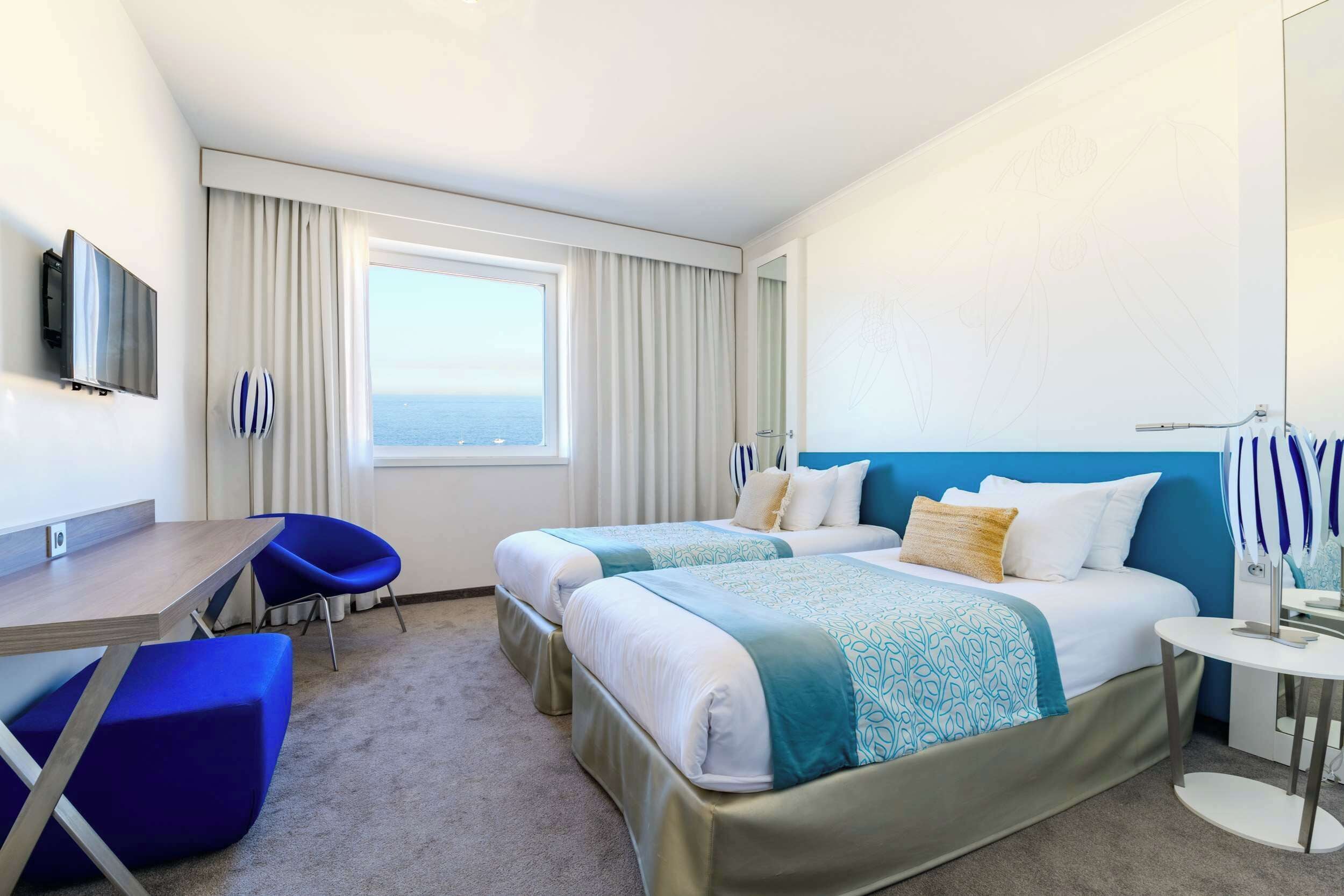 A hotel room with two beds, a desk, a chair, a TV, and a view of the sea.