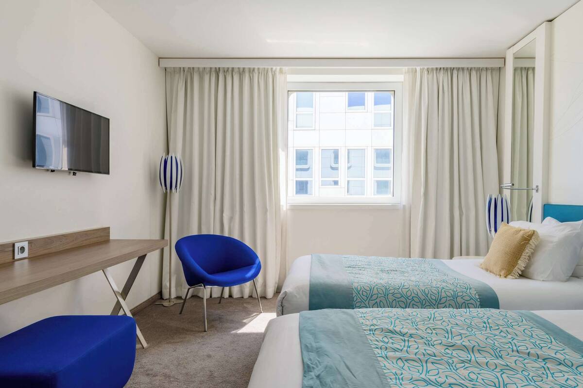 A hotel room with a bed, a desk, a chair, a TV, and a window with curtains.