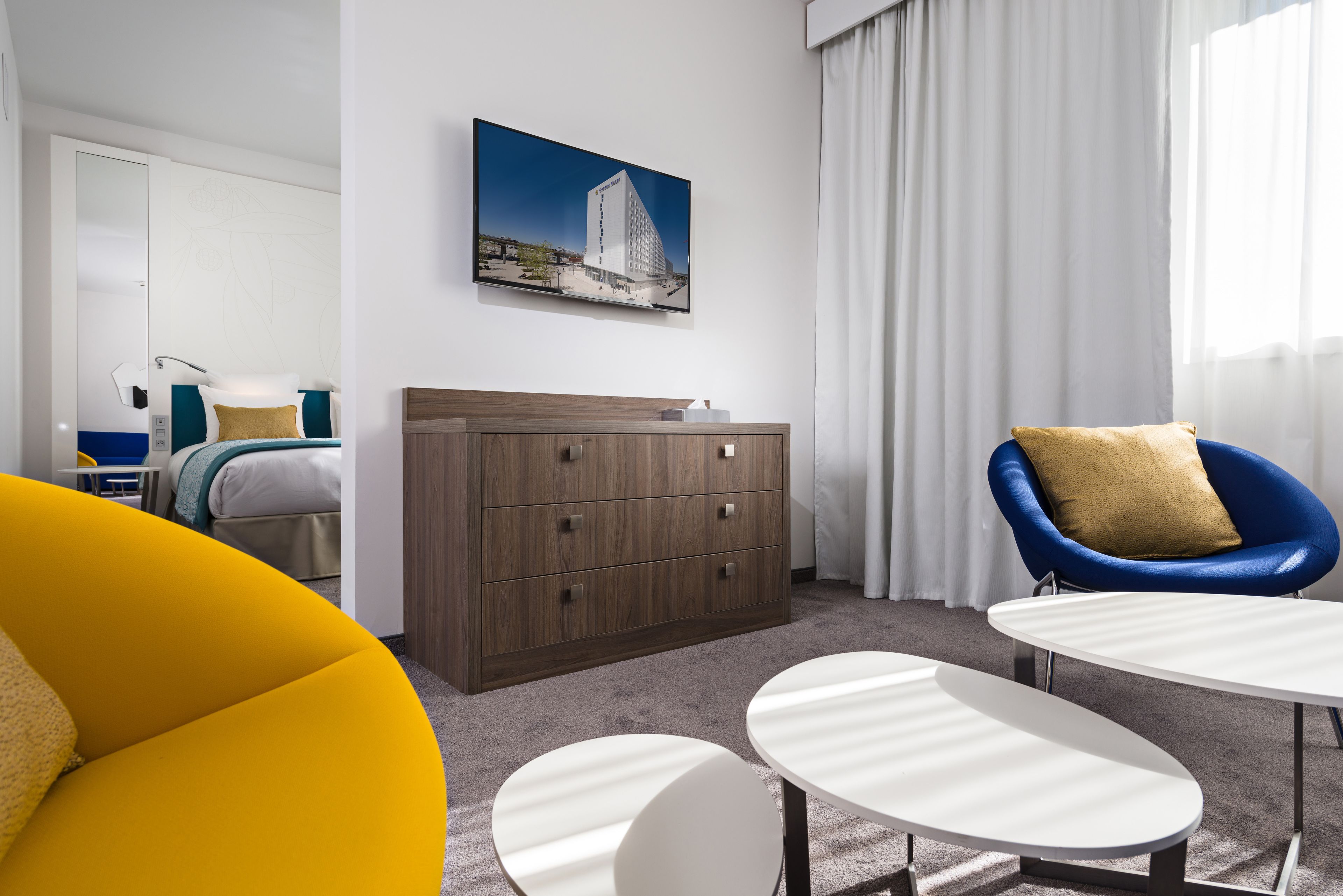 Una habitación de hotel moderna con una cama, un aparador de madera, un sillón azul y una pequeña mesa blanca.