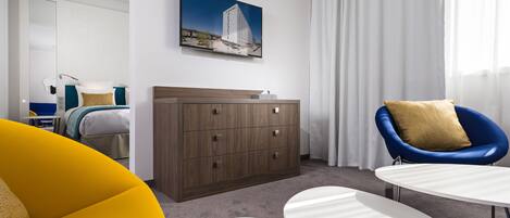 Una habitación de hotel moderna con una cama, un aparador de madera, un sillón azul y una pequeña mesa blanca.