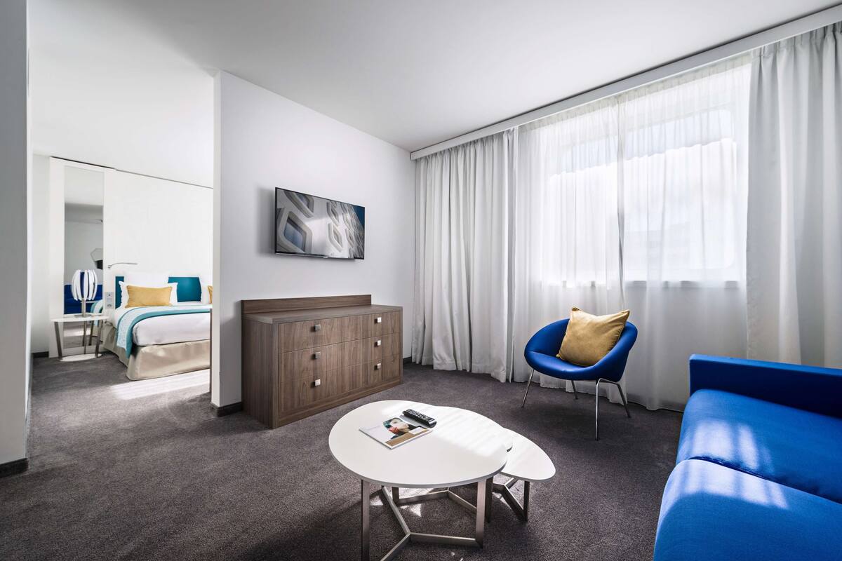 A modern hotel room with a bed, a white coffee table, a blue sofa, a wooden cabinet, and a large window with sheer curtains.