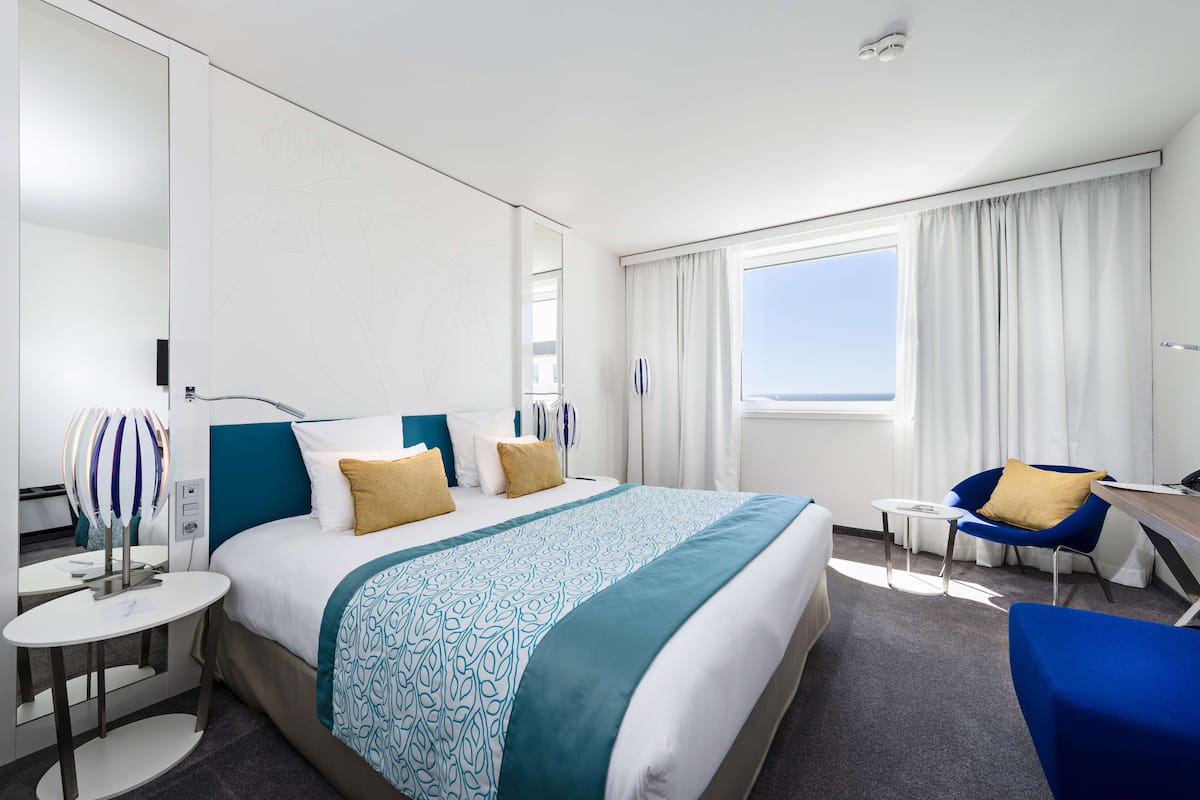 A hotel room with a large bed, a desk, a chair, and a window with curtains.