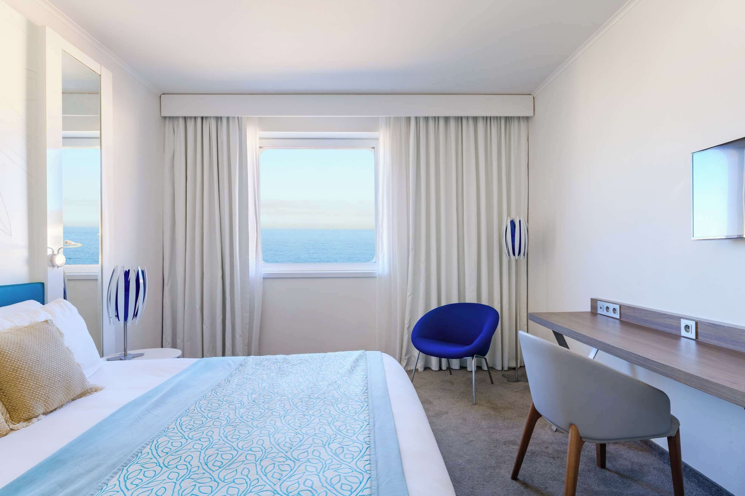 Habitación de hotel con cama, escritorio, silla, ventana grande con cortinas y vista al mar.