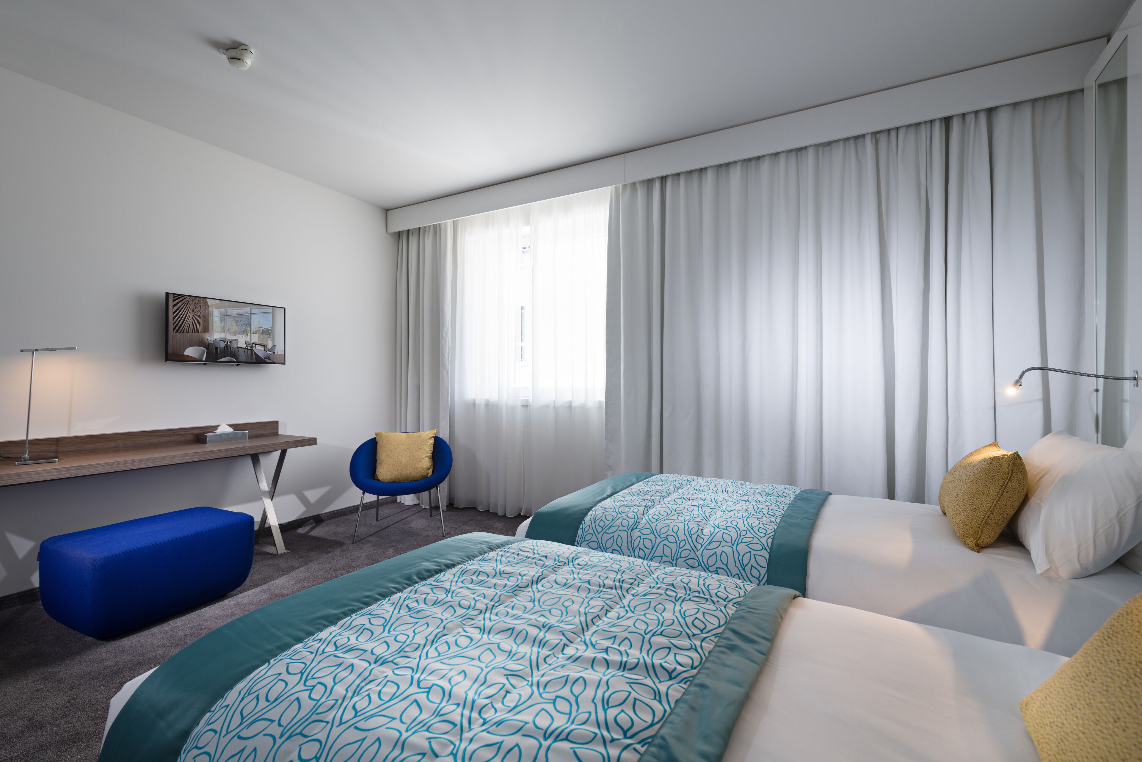 Camera d'albergo con due letti, una scrivania, una sedia e un'ampia finestra con tende.