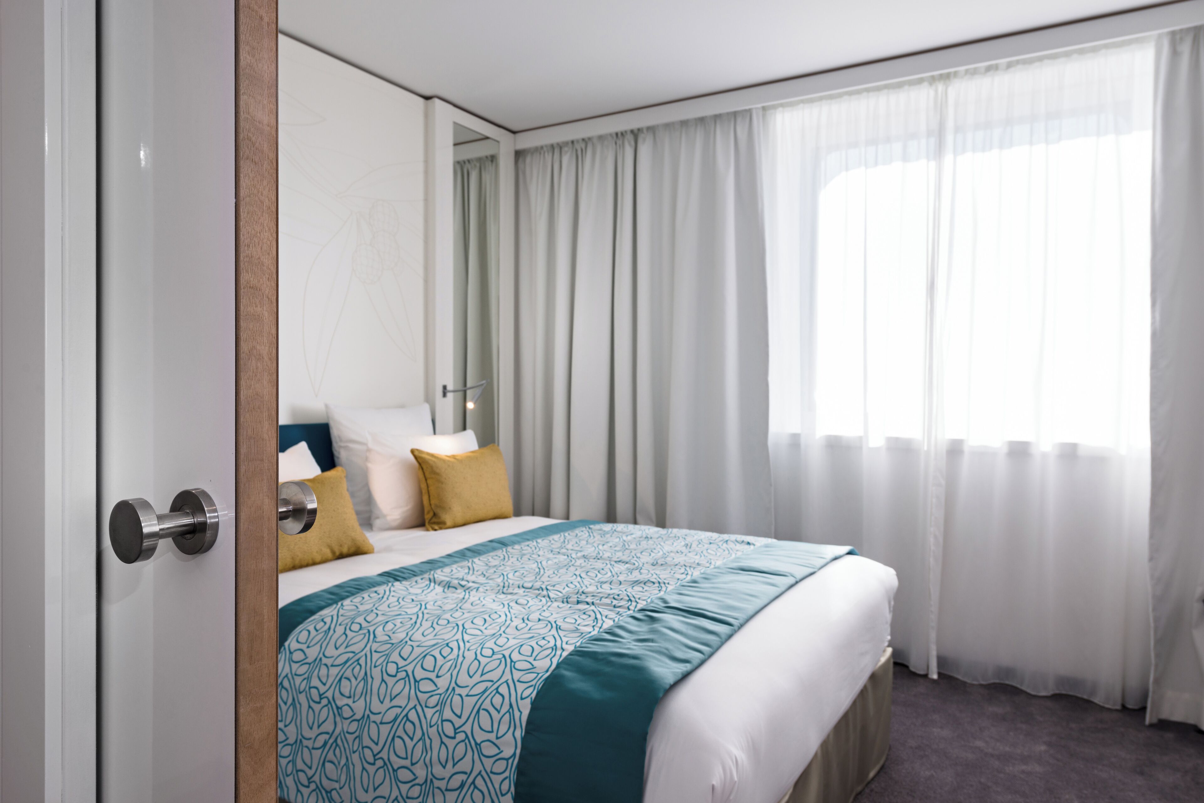 Quarto de hotel com uma cama grande, porta de madeira e janela com cortinas leves.