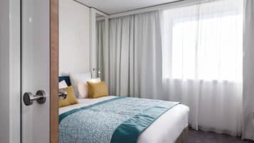 Quarto de hotel com uma cama grande, porta de madeira e janela com cortinas leves.