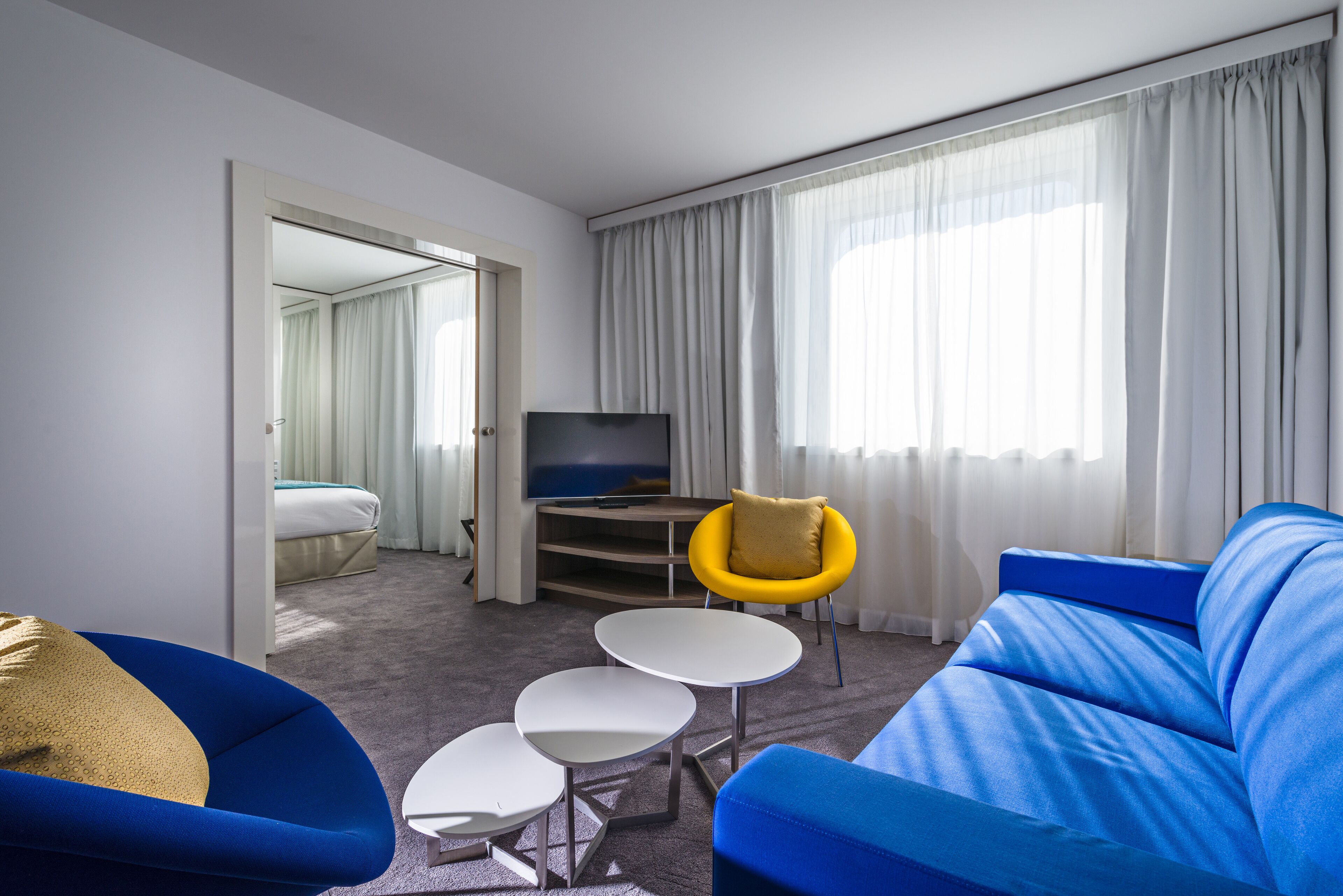 Ein modernes Hotelzimmer mit einem blauen Sofa, einem gelben Sessel, einem Flachbildfernseher und einem großen Fenster mit Vorhängen.