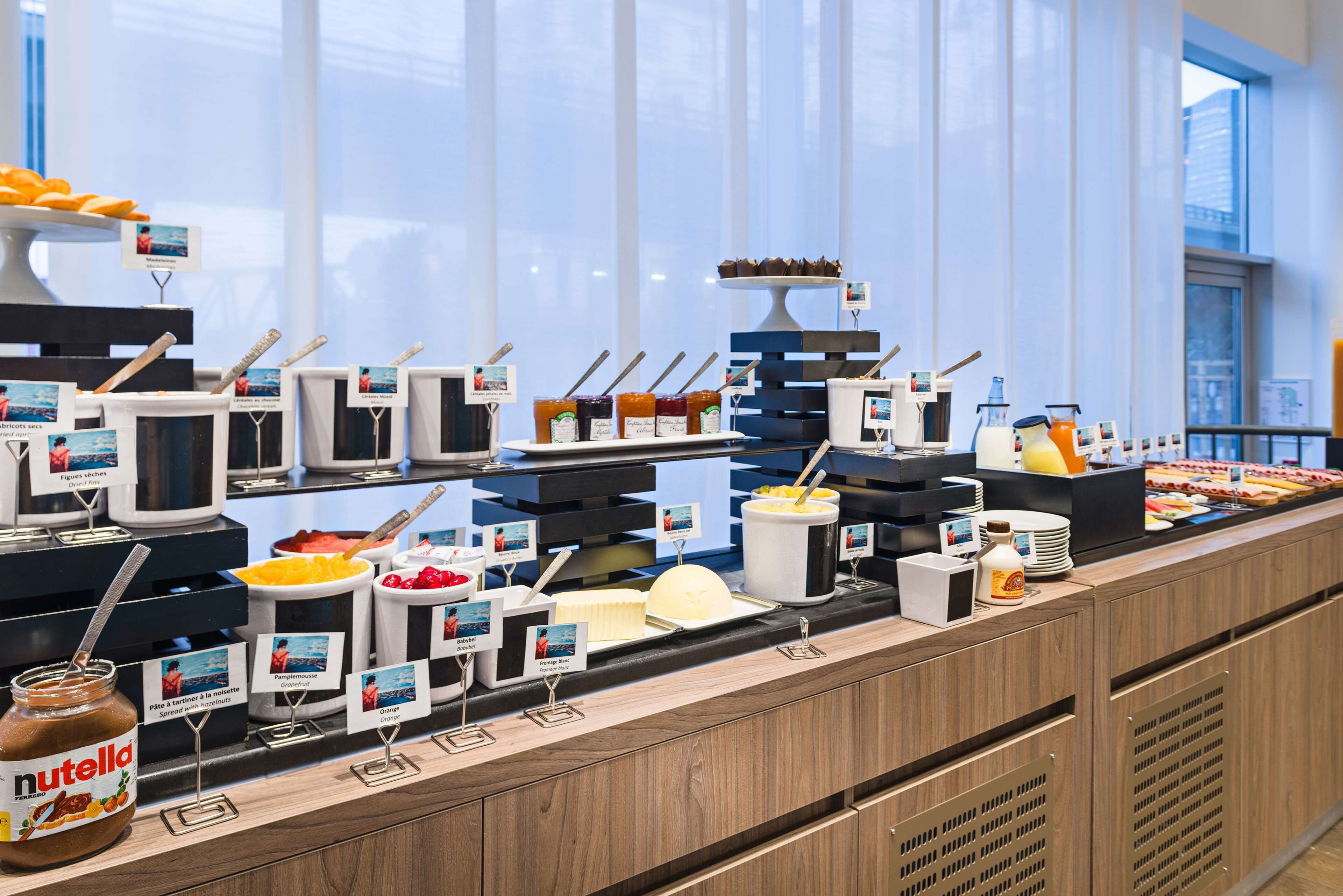 Buffet com diversos alimentos e acompanhamentos, incluindo potes de Nutella e outras pastas, disposto em um balcão de madeira com vista para a cidade ao fundo.