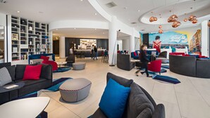 Um hall de hotel moderno com um bar central, áreas de estar e uma grande estante de livros.
