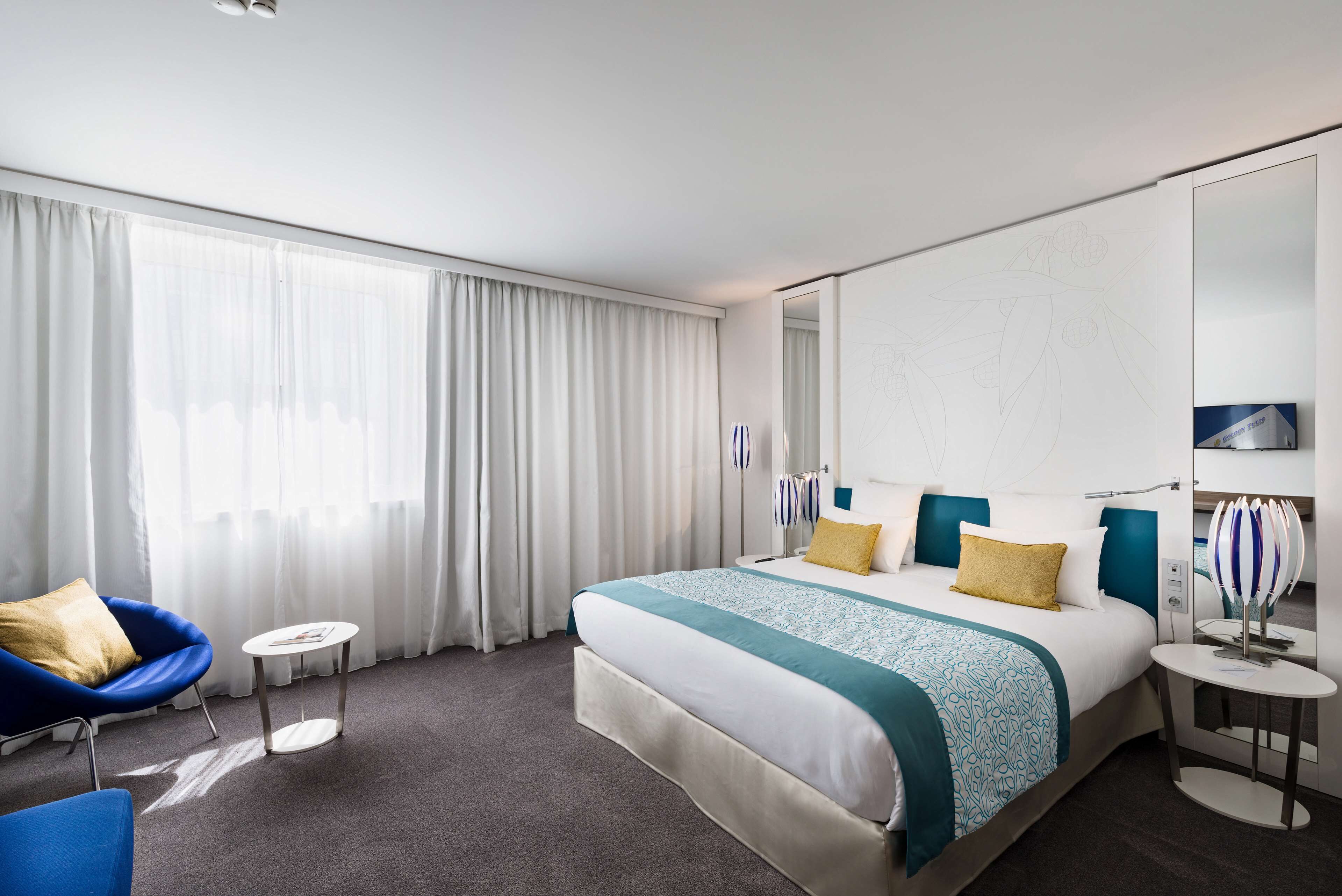Une chambre d’hôtel moderne avec un grand lit, une chaise bleue, une petite table et une grande fenêtre avec des rideaux.