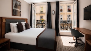 A hotel room with a bed, a desk, a chair, a TV, and a balcony with a view of buildings. - Mayflower Opéra  (Paris)